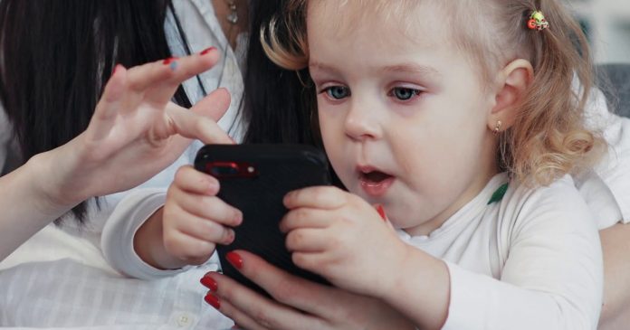 videoblocks-mom-and-daughter-sit-on-sofa-use-phone-hug-woman-baby-hand-love-child-person-girl-kid-internet-cartoon-technology-smartphone-face-touchscreen-home-mobile-looking-application-tou